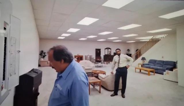 A lower level furniture showroom with a breaker box in the foreground, water stained ceiling tiles, and a staircase.